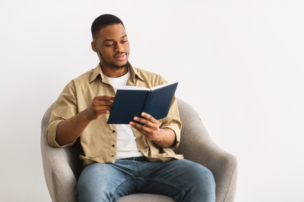 Afrikanischer Kerl liest Buch auf Stuhl über grauem Hintergrund - Foto, Bild