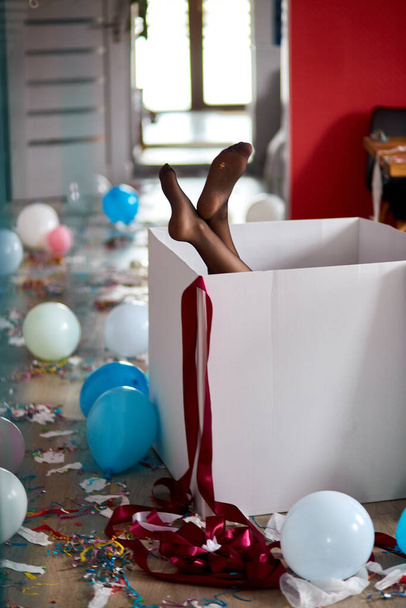 Woman legs sticking from big box with leaky stockings, after party chaos, messy in living room at home, confetti and balloons, morning after party celebration. - Fotoğraf, Görsel