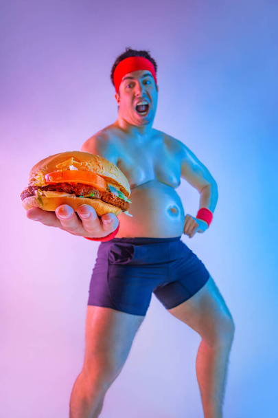 Fat man with gives away hamburger for lose weight and become a slim athlete. Not sporty male eating hamburger isolated on pink background. - 写真・画像