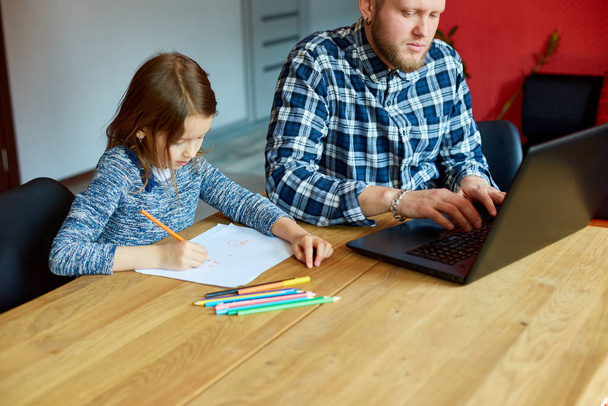 Батько працює в своєму домашньому офісі на ноутбуці, її дочка сидить поруч з нею і малює, чоловік позаштатний, віддалена робота і виховання дитини на робочому місці
. - Фото, зображення