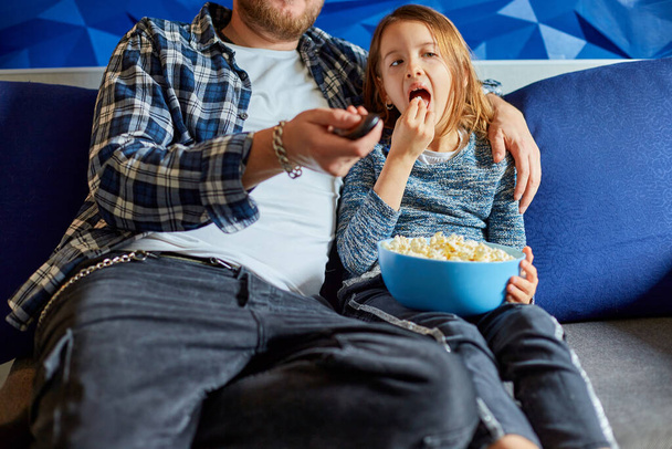 Vater und Tochter essen Popcorn und gucken Fernsehfilm, Vater und Kind schauen Film auf der Couch zu Hause, glücklich lächelnde Familie verbringt Freizeit zusammen, entspannen. - Foto, Bild