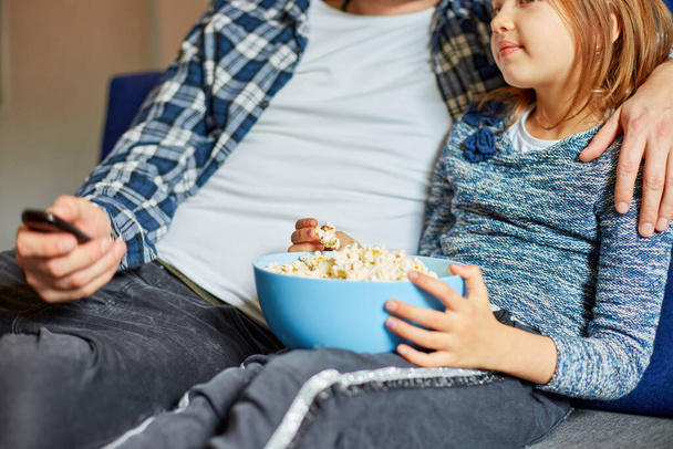 Father and daughter eat popcorn and watch TV film, Dad and child girl watching a movie on a couch at home, happy smiling family spending free time together, relax. Close up hands, - Photo, Image