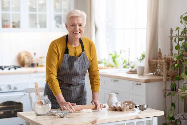 Ελκυστική ηλικιωμένη γυναίκα κάνει ζύμη στην κουζίνα, χρησιμοποιώντας πλάστη - Φωτογραφία, εικόνα