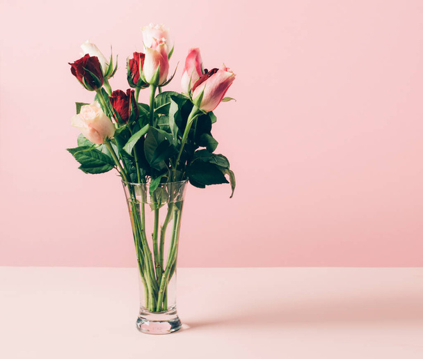 Красивые свежие розы в стеклянной вазе расположены на столе на пастельно-розовом фоне. Выборочное пространство для копирования фокуса - Фото, изображение
