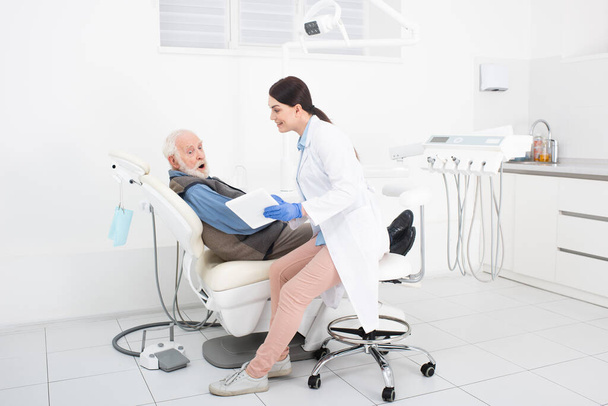 paciente sênior espantado deitado na cadeira dentária e tendo consulta do médico com tablet nas mãos - Foto, Imagem