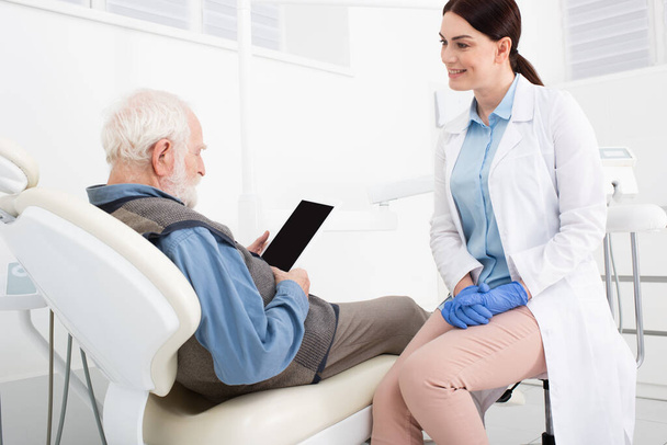 高齢の患者は歯の椅子に横になり診療所の医師の近くに黒い画面のタブレットを持っています - 写真・画像