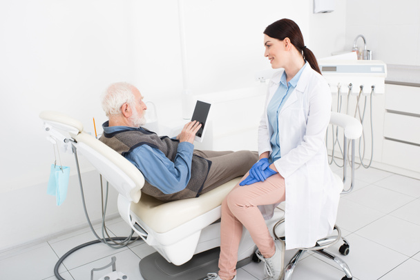 senior man lying in dental chair and looking at tablet near doctor in dental clinic - Фото, изображение
