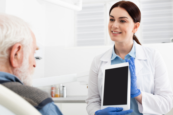 vrolijke tandarts tonen tablet aan senior patiënt in tandheelkundige kliniek - Foto, afbeelding