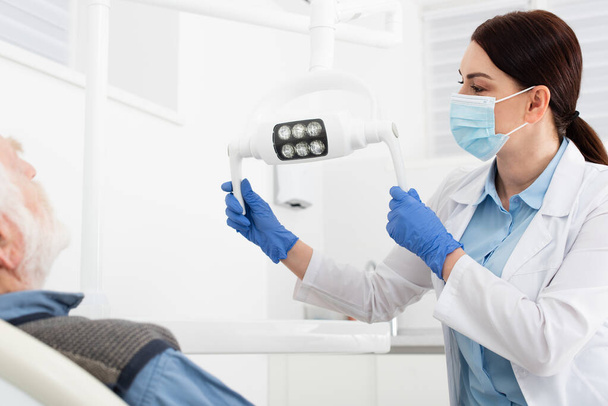 dentista en máscara médica y guantes de látex azul tocando lámpara médica delante de paciente mayor - Foto, imagen