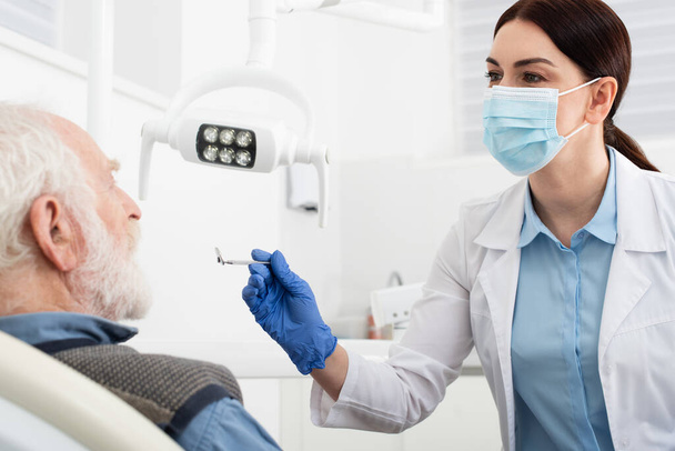 пожилой человек с осмотром зубов стоматологом в латексных перчатках с зеркалом в стоматологическом кресле - Фото, изображение