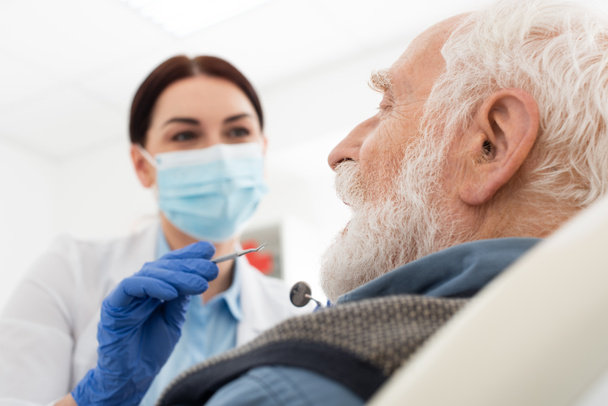 歯科用椅子のプローブとミラーで老人の歯を調べる医療用マスクの歯科医 - 写真・画像
