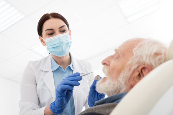 стоматолог в медицинской маске, осматривающий зубы пожилого человека с зондом и зеркалом в стоматологической клинике - Фото, изображение