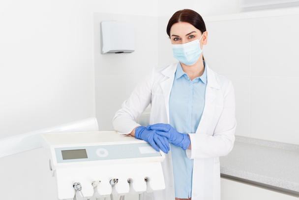 zubař v latexových rukavicích a lékařské masce stojí v blízkosti zařízení a dívá se na kameru v zubní klinice - Fotografie, Obrázek