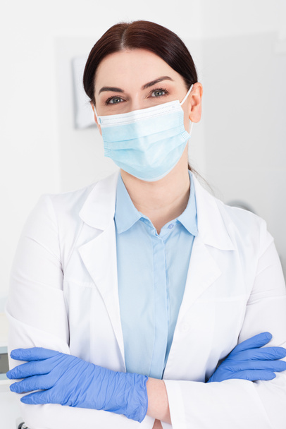 portret van tandarts in medisch masker en latex handschoenen staande met gekruiste armen in tandheelkundige kliniek - Foto, afbeelding