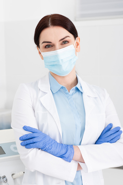dentista en guantes de látex y máscara médica de pie con los brazos cruzados en la clínica dental - Foto, imagen