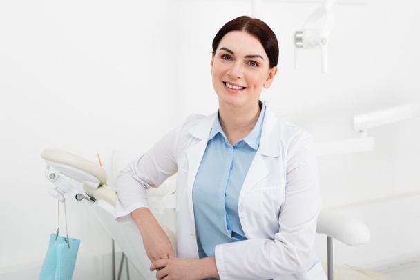 portrét veselého zubaře v bílém plášti sedícího na zubní klinice  - Fotografie, Obrázek