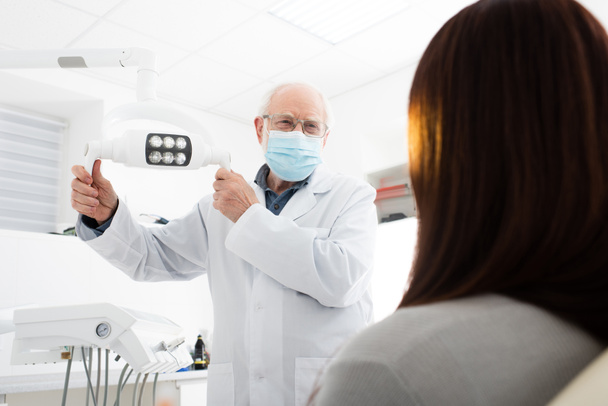 dentista sênior em máscara médica tocando lâmpada médica na frente do paciente  - Foto, Imagem