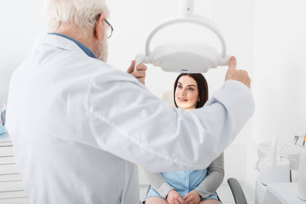 senior dentist regulating medical lamp in front of patient in dental chair - Photo, Image
