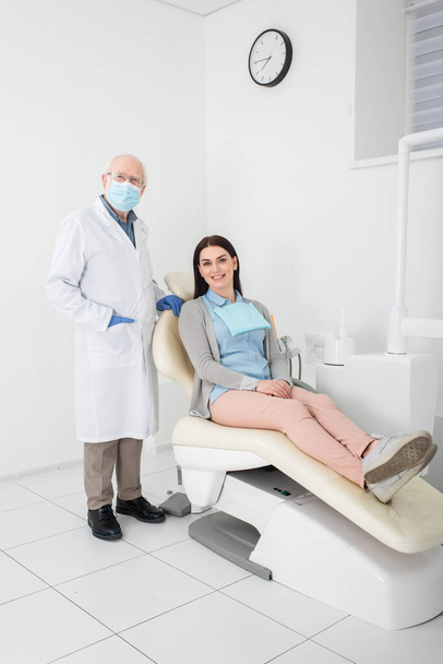 starší zubař v bílém plášti, lékařské masce a latexových rukavicích stojící poblíž pacienta v zubním křesle - Fotografie, Obrázek