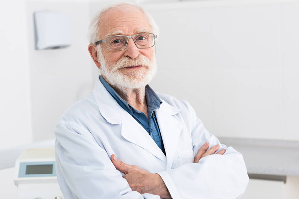 portrait of senior dentist in white coat and glasses standing with crossed arms in dental clinic - Фото, зображення