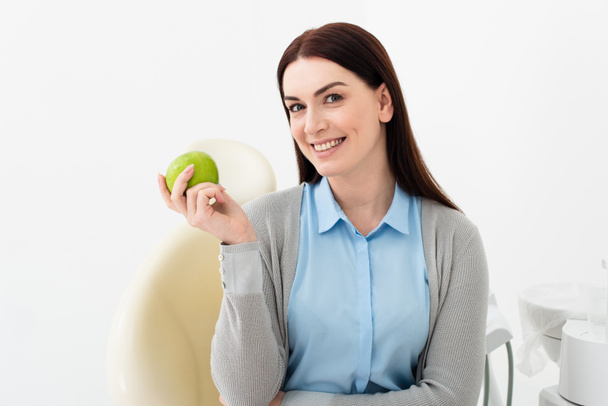 mulher adulta sorridente na cadeira dental segurando maçã verde e olhando para a câmera na mão na clínica - Foto, Imagem