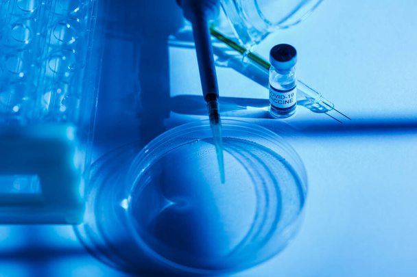 Mesa de laboratorio con soporte para tubos de ensayo, vial con vacuna para COVID-19, jeringa, pipeta, gafas protectoras y placa petri, con ambiente de luz azul - Foto, Imagen