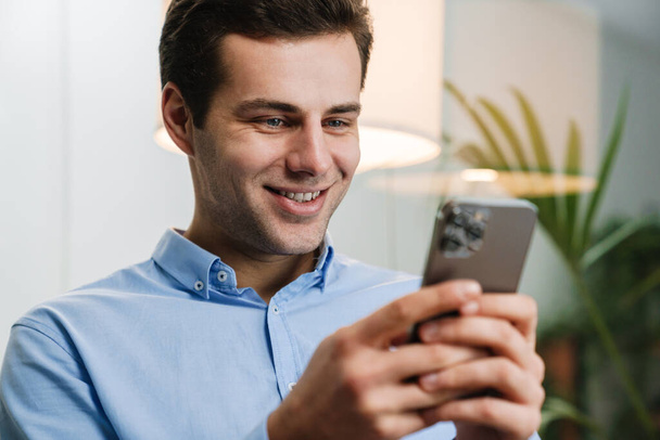 Smiling handsome young entrepreneur using mobile phone while sitting in a modern office - Fotoğraf, Görsel