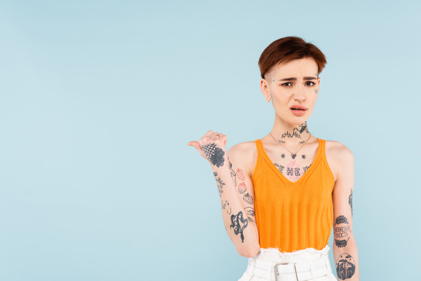 displeased and tattooed woman pointing with thumb away isolated on blue  - Photo, Image