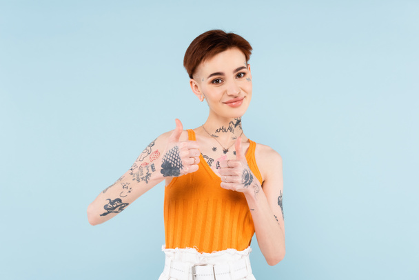 pleased and tattooed woman showing thumbs up isolated on blue  - Fotoğraf, Görsel