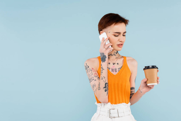 mujer tatuada hablando en el teléfono inteligente mientras sostiene el café para ir aislado en azul  - Foto, Imagen