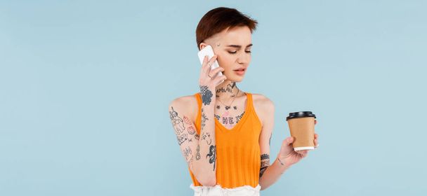 tattooed woman talking on smartphone while holding coffee to go isolated on blue, banner - Photo, image