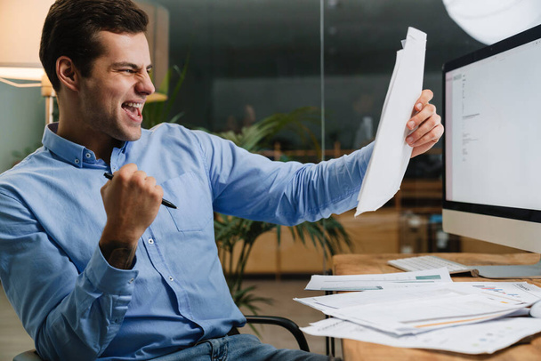 Happy cheerful young man celebrating success while sitting in the office with paperwork - Fotoğraf, Görsel