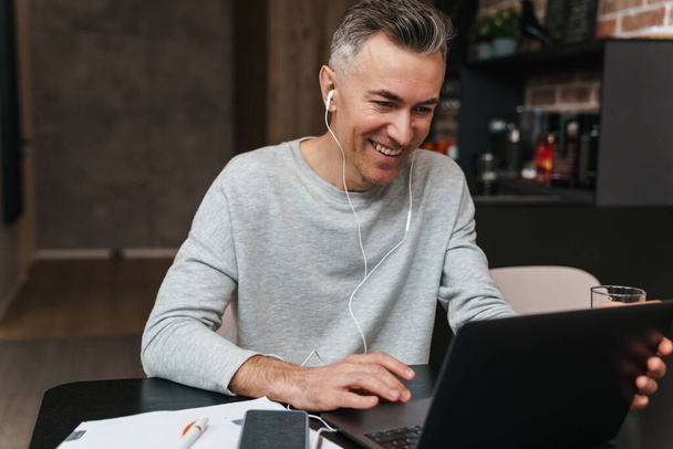 Handsome smiling young man on a video conference via laptop computer, wearing headphones, sitting at the desk with paper work - Фото, изображение