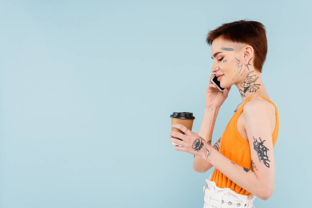 side view of smiling and tattooed woman talking on smartphone while holding paper cup isolated on blue - Foto, Imagem