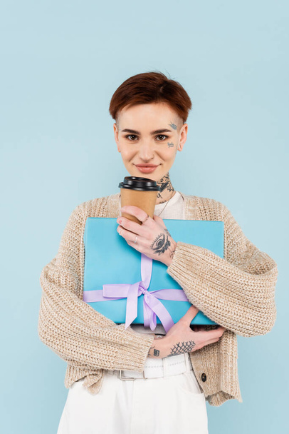Gülümseyen dövmeli kadın, elinde hediye paketi ve mavi bardakta izole edilmiş kağıt bardak tutuyor.  - Fotoğraf, Görsel