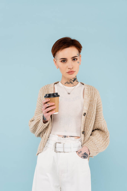 young tattooed woman holding paper cup while standing with hand in pocket isolated on blue  - Photo, image