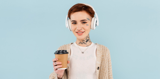 smiling tattooed woman in wireless headphones holding paper cup isolated on blue, banner - Foto, imagen