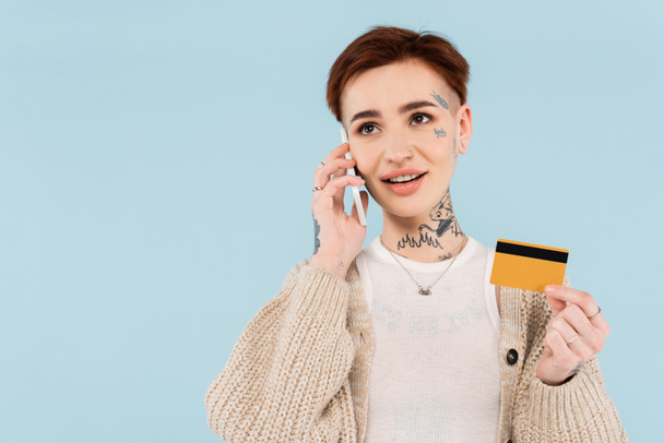 mujer alegre y tatuada sosteniendo la tarjeta de crédito mientras que hace el orden en el teléfono inteligente aislado en azul - Foto, imagen
