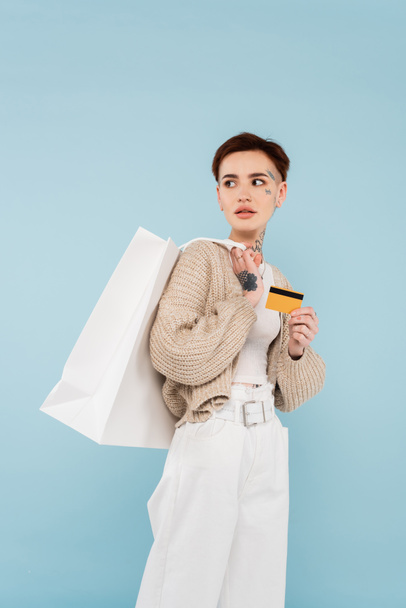 young woman with tattoos holding credit card and shopping bags while looking away isolated on blue - Foto, afbeelding