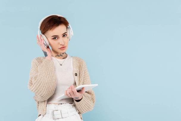 young woman with tattoos holding digital tablet and listening music in wireless headphones isolated on blue - Foto, imagen