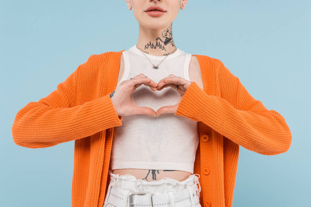 καλλιεργημένη άποψη της νεαρής γυναίκας με τατουάζ σε πορτοκαλί ζακέτα που δείχνει την καρδιά με τα χέρια απομονώνονται σε μπλε - Φωτογραφία, εικόνα