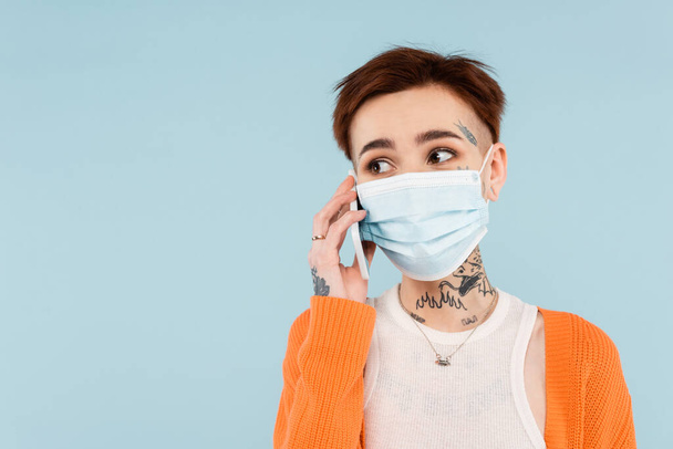 tattooed woman in orange cardigan and medical mask talking on smartphone isolated on blue - Foto, immagini