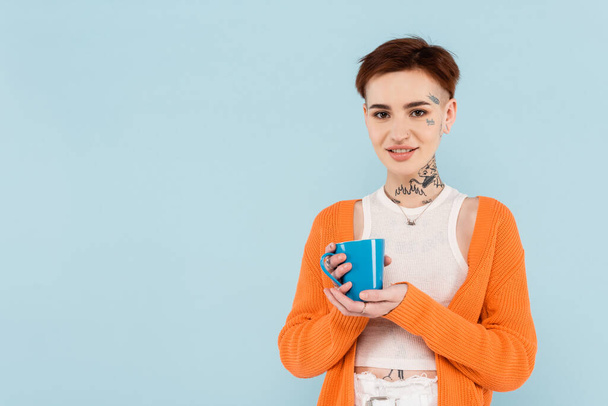 happy tattooed woman in orange cardigan holding cup of coffee isolated on blue - Zdjęcie, obraz