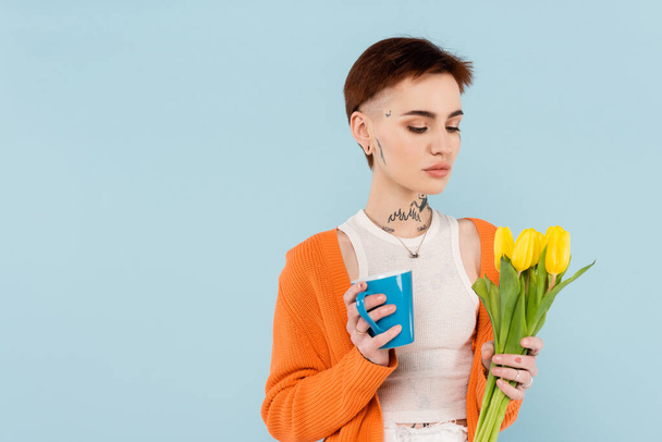 young tattooed woman in orange cardigan holding cup of coffee and yellow tulips isolated on blue - Fotografie, Obrázek