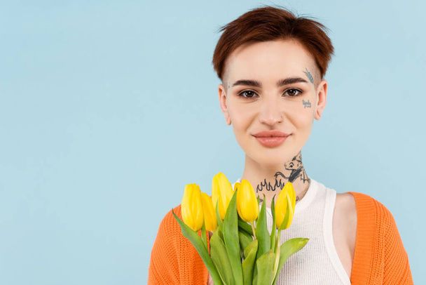 cheerful tattooed woman in orange cardigan holding yellow tulips isolated on blue - Foto, Imagen