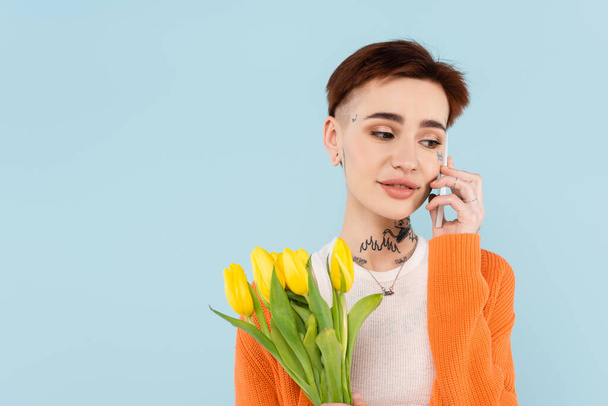 young tattooed woman in orange cardigan holding tulips while talking on smartphone isolated on blue - Foto, Imagem