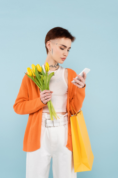 young woman with tattoos holding yellow tulips and shopping bag while chatting on cellphone isolated on blue - Fotografie, Obrázek