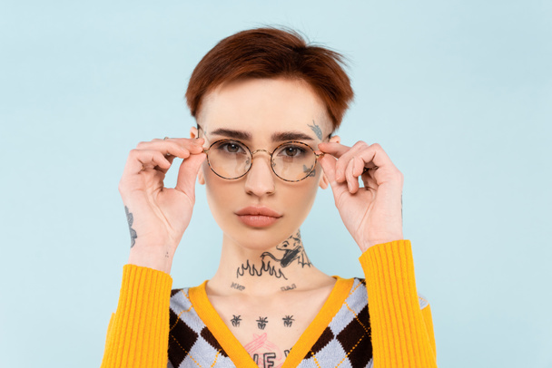 mujer joven tatuada ajustando gafas aisladas en azul - Foto, Imagen