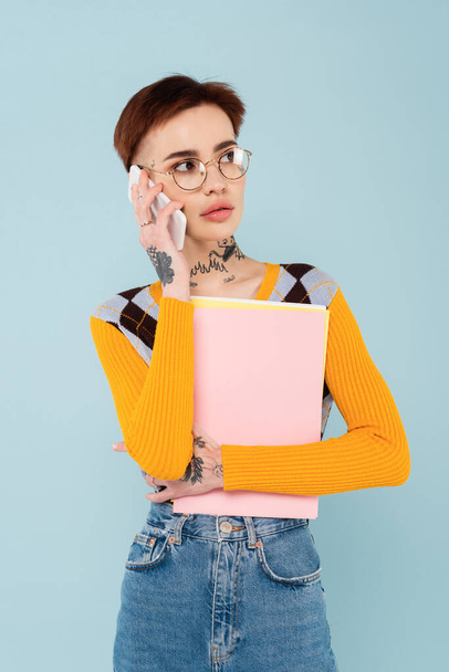 young tattooed student in glasses talking on cellphone and holding folder isolated on blue - Photo, Image