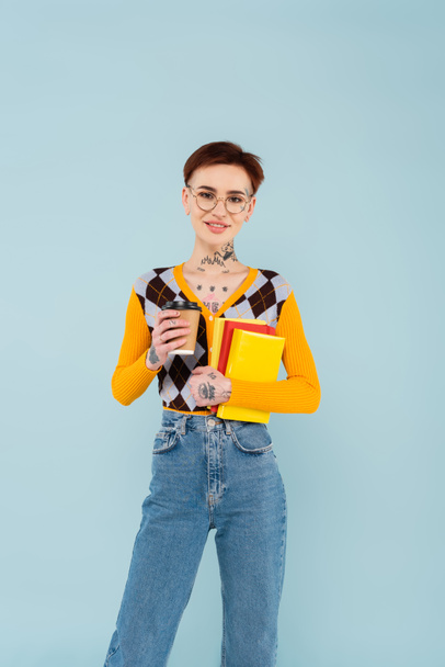 happy tattooed student in glasses holding books and paper cup isolated on blue - Фото, изображение